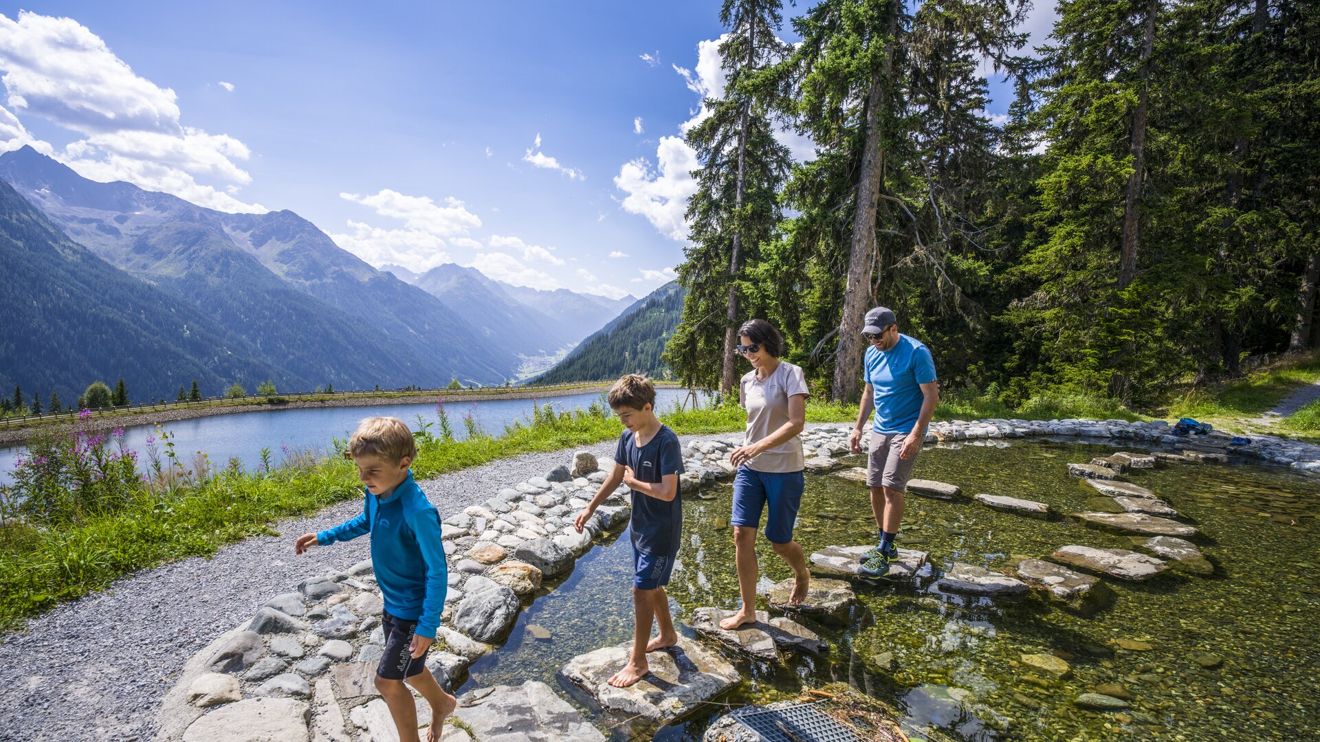 Eine Familie mit zwei Buben balanciert über einen Steinpfad in einem flachen Kneippbecken mitten im Sunny Mountain Erlebnispark.
