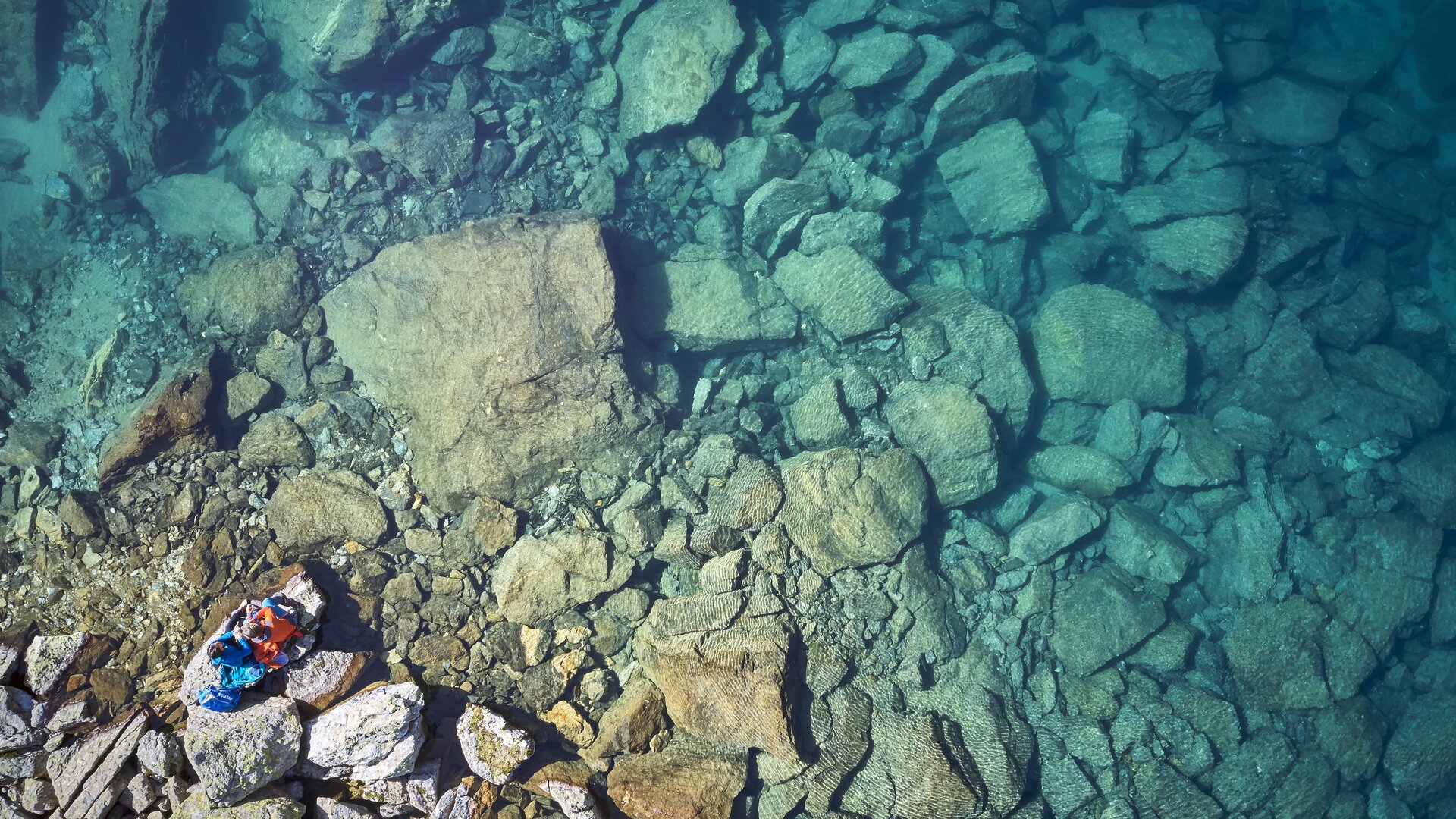 Ein klarer Bergsee in Kappl aus der Vogelperspektive. Am Grund des Sees sind große Steine und Felsen zu erkennen.