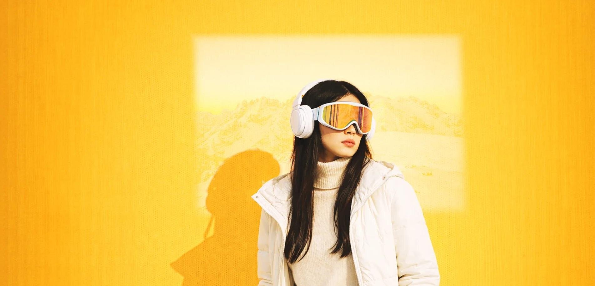Eine Frau steht vor einer gelben Wand und trägt eine weiße Jacke sowie farbige Skibrille. Im Hintergrund ist eine verschwommene Landschaft zu sehen.
