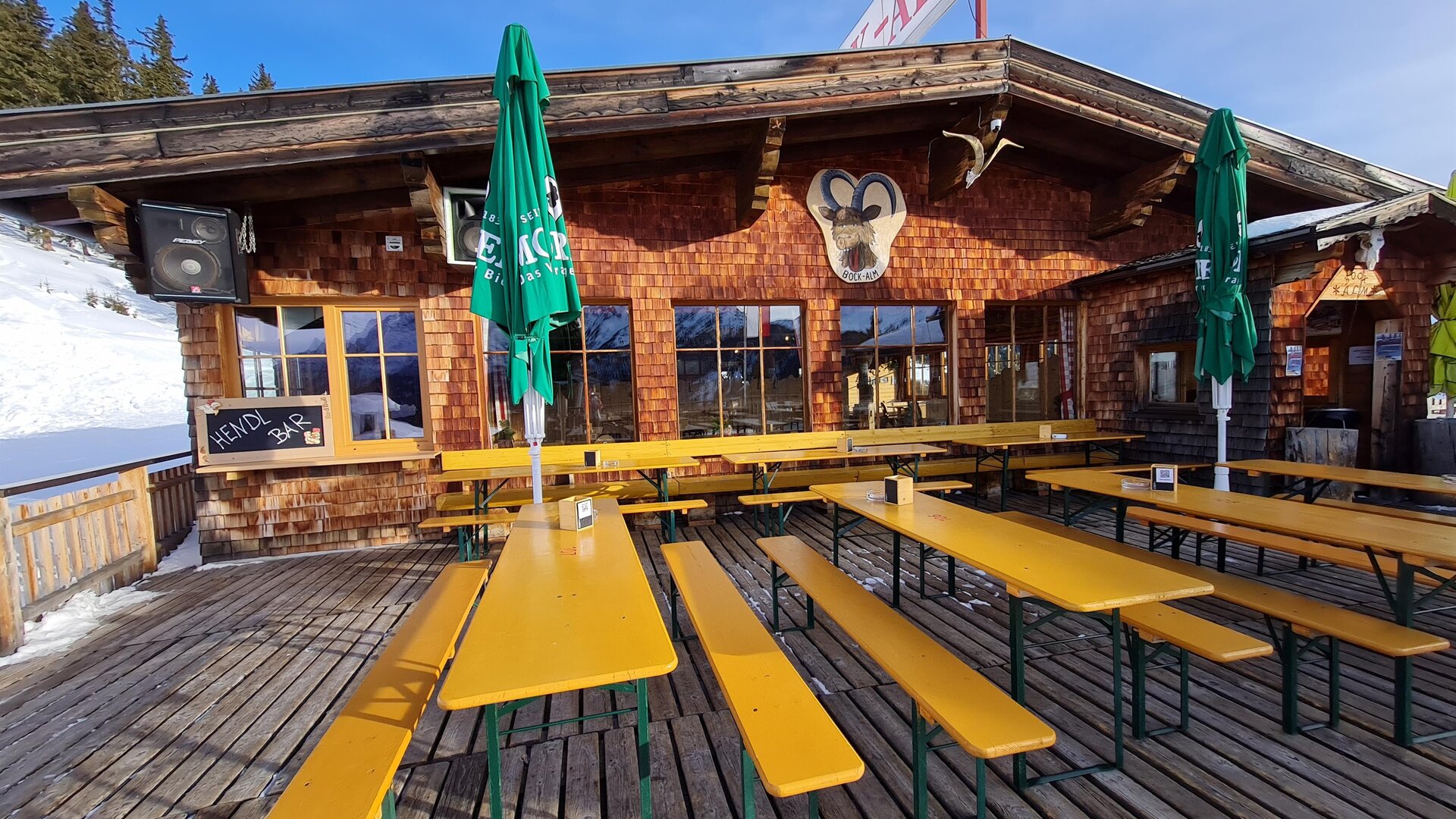 A cozy mountain cabin with wooden cladding and several long benches. The terrace is equipped with green sun umbrellas and the snow is visible in the background.