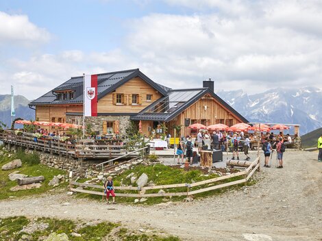 A cozy mountain cabin with a terrace and many visitors. In the background, high mountains and a partly cloudy sky can be seen.