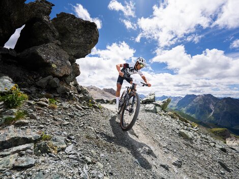 A mountain biker rides on a rocky path in the mountains. In the background, impressive mountain landscapes and a blue sky with clouds are visible.