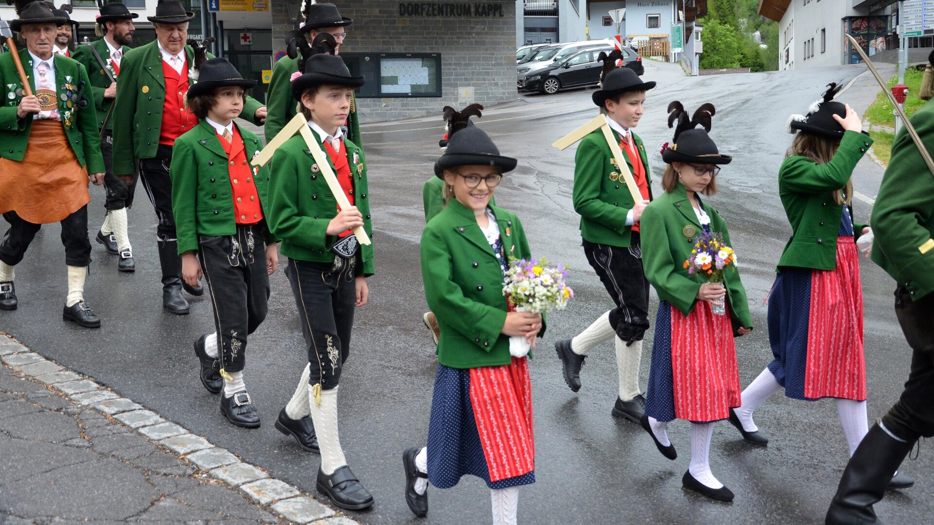 A group of traditional costume bearers marches through a street. They wear green jackets and black hats, and some hold flowers in their hands.