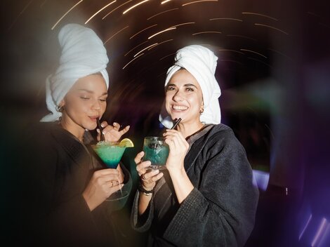 Two women in bathrobes and with towels on their heads are holding colorful cocktails. In the background, lights and a relaxed atmosphere can be seen.