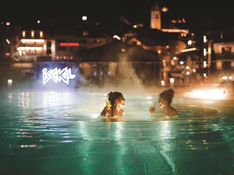 Zwei Frauen entspannen im beleuchteten Pool bei Nacht. Im Hintergrund sind die Lichter der Stadt sichtbar.
