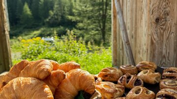 A basket full of freshly baked croissants is next to a plate with various pastries. In the background, green trees and mountains are visible.