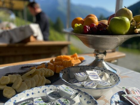 A beautifully set table with various foods, including fresh fruit and portions of cream cheese. In the background, clear mountains and a blue sky can be seen.