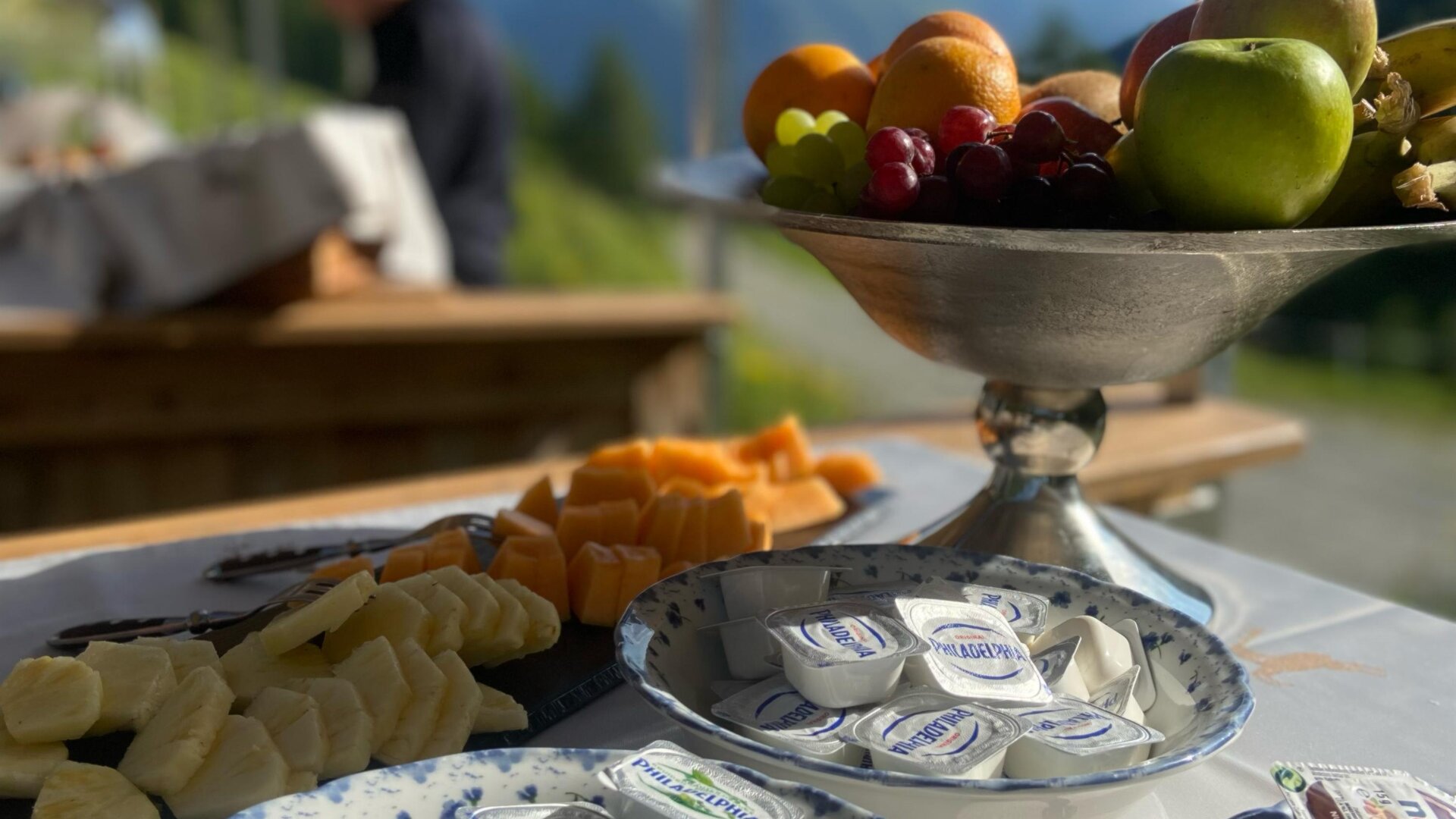 A beautifully set table with various foods, including fresh fruit and portions of cream cheese. In the background, clear mountains and a blue sky can be seen.