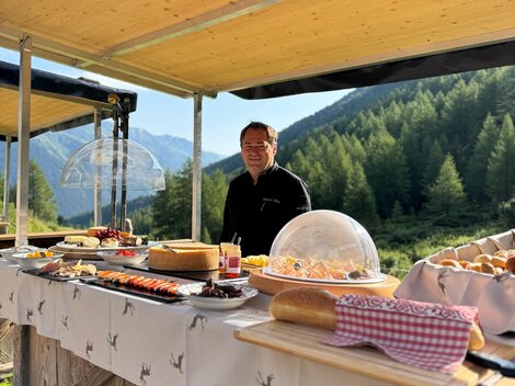 A friendly vendor stands at a wooden stand with a variety of foods. In the background, mountains and forests can be seen.