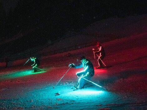 Three skiers with bright, colorful lights descend a slope in the dark. The surroundings are covered in snow and the atmosphere is festive.