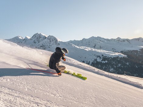 Ein Skifahrer fährt über eine schneebedeckte Piste in den Bergen. Im Hintergrund sind majestätische Berge und ein klarer Himmel zu sehen.