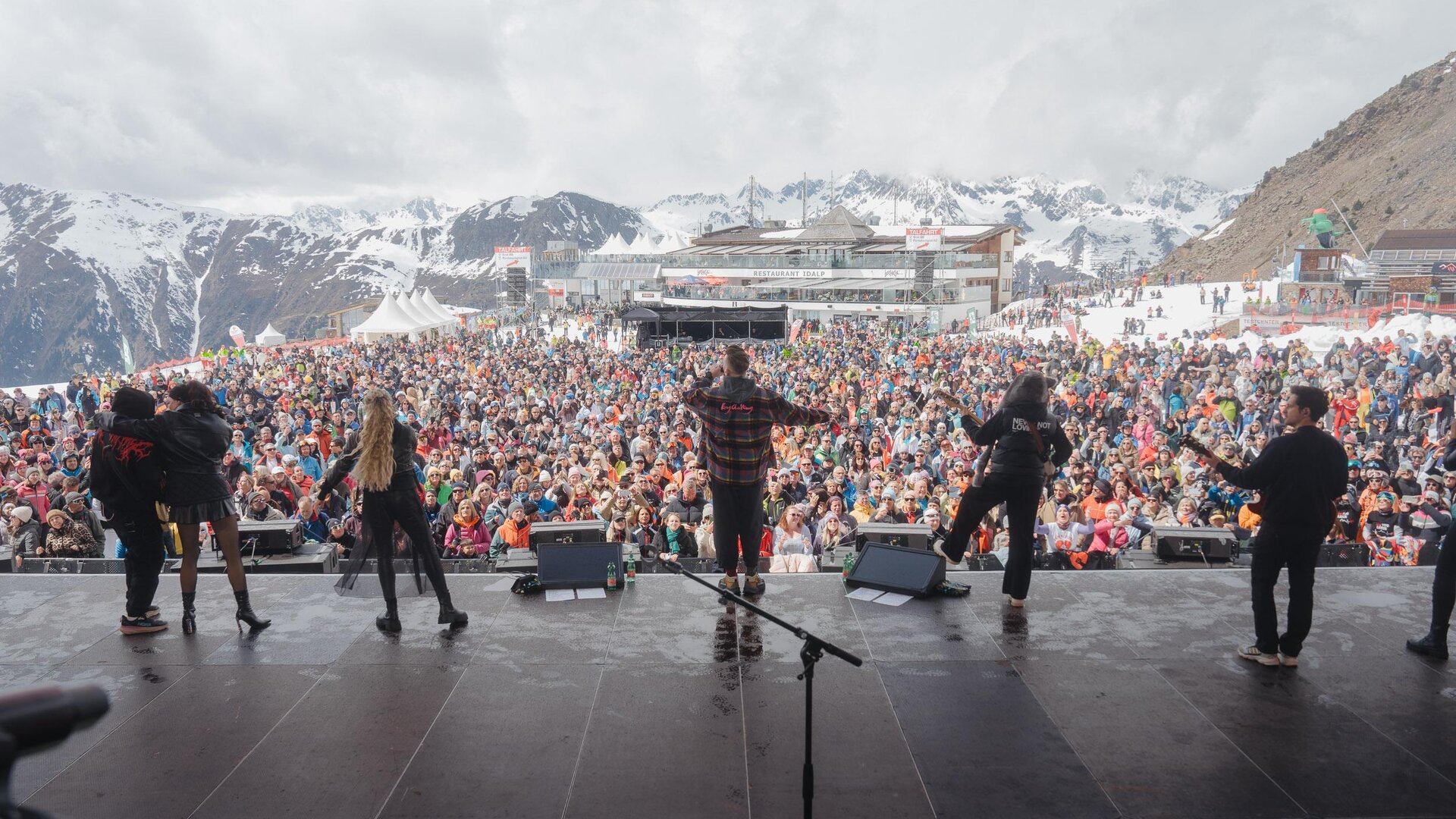 A large crowd enjoys a concert in the mountains. In the background, there are snow-covered peaks and an event building.