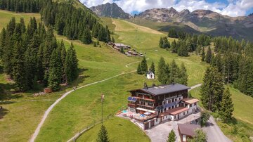 Ein malerisches Bergpanorama mit viel Grün und Wolken am Himmel. Im Vordergrund steht ein gemütliches Hotel umgeben von Wälder und Wanderwegen.