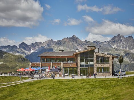 A modern building in a picturesque mountain landscape. The terrace is adorned with guests and colorful umbrellas.