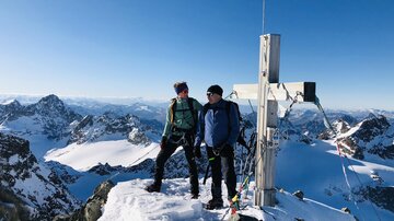 Zwei Bergsteiger stehen auf einem schneebedeckten Gipfel. Im Hintergrund sind beeindruckende Berge und ein klarer blauer Himmel zu sehen.