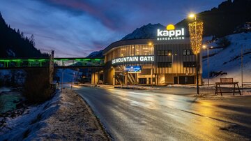 Ein modernes Gebäude mit dem Schriftzug "Kappl Bergbahnen" steht im Vordergrund. Die Szenerie zeigt eine winterliche Umgebung bei Dämmerlicht.