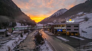 Ein malerischer Blick auf eine winterliche Landschaft mit schneebedeckten Bergen und einem Fluss. Im Vordergrund steht das Gebäude "Mountain Gate" mit einem farbenfrohen Sonnenuntergang im Hintergrund.
