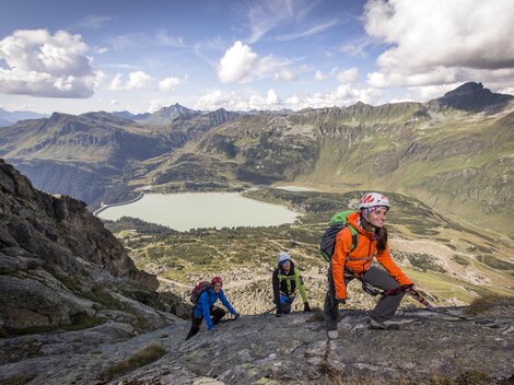 A group of climbers is ascending a rock in the Alps. In the background, a clear lake and impressive mountain landscapes can be seen.