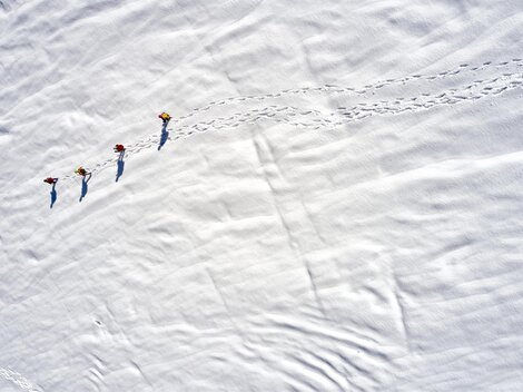 Four people are walking across a snow-covered area. Their tracks are clearly visible in the fresh snow.