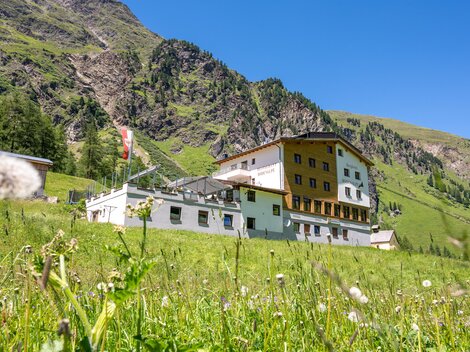 A large building in the mountains, surrounded by green meadows and rocks. The sky is clear and blue.