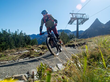 A cyclist is riding on a narrow path in the mountains. In the background, the mountain peaks and a cable car can be seen.