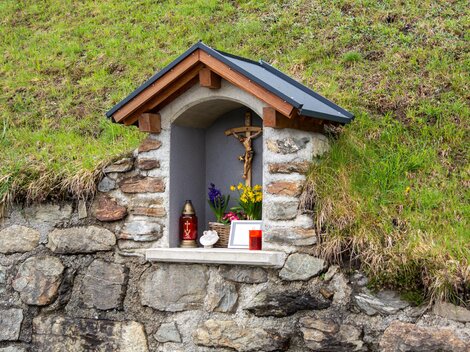 Eine kleine Nische in einer Steinmauer mit einem Holzdach. Darin befinden sich Figuren, Kerzen und Blumen zur Andacht. | © Bernhard Gruber – Photography