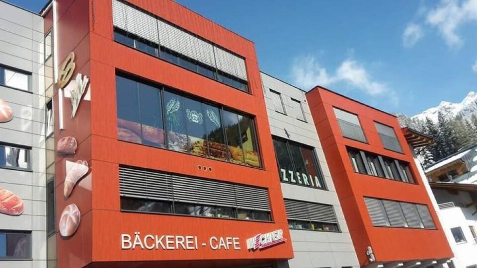 Ein modernes Gebäude mit roter Fassade und großen Fenstern. Darin sind ein Bäckerei-Café und eine Pizzeria angesiedelt.