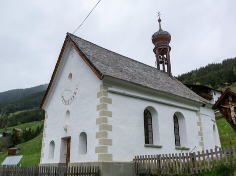 Eine kleine, weiße Kirche mit einem hohen Turm steht in einer grünen Landschaft. Im Hintergrund sind sanfte Hügel und Wälder zu sehen. | © Bernhard Gruber – Photography