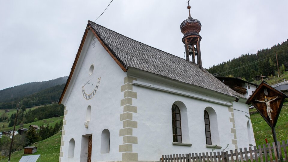 Eine kleine, weiße Kirche mit einem hohen Turm steht in einer grünen Landschaft. Im Hintergrund sind sanfte Hügel und Wälder zu sehen. | © Bernhard Gruber – Photography