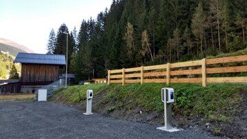 A rural setting with a wooden building and a row of fences. In the foreground, there are two charging stations for electric vehicles.