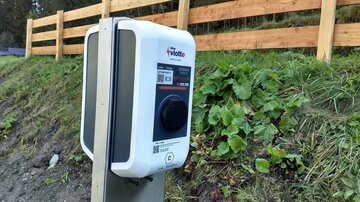 An electric vehicle charging station is located next to a green meadow. In the background, a wooden fence and trees can be seen.