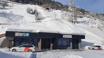 Ein Geschäft in einer verschneiten Landschaft mit Bäumen und hügeligem Gelände. Die Umgebung ist winterlich und üppig mit frischem Schnee bedeckt.