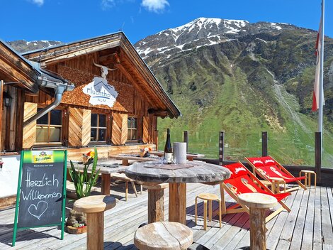 A cozy mountain cabin with wooden elements and a magnificent mountain landscape in the background. There is an audience outside with lounge chairs and a welcome sign.