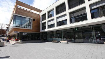 Ein modernes Gebäude mit großen Fenstern und Holzverkleidungen. Der Platz davor ist gepflastert und bietet Sitzmöglichkeiten.