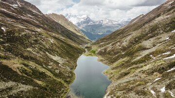 Eine idyllische Berglandschaft mit einem klaren See in der Mitte. Umgeben von grünen Hügeln und majestätischen Bergen unter einem bewölkten Himmel.