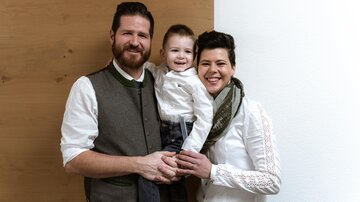 A family with a small child stands together in front of a bright wall. They are all wearing elegant, casual clothing and smiling kindly.