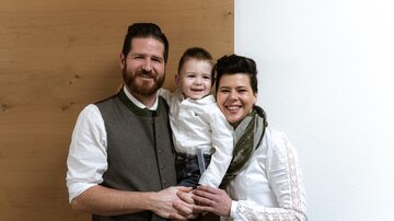 A family with a small child stands together in front of a bright wall. They are all wearing elegant, casual clothing and smiling kindly.