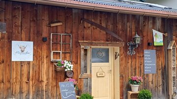 Ein rustikales Holzgebäude mit einer einladenden Eingangstür. Blumen und Pflanzen schmücken den Eingangsbereich, und eine Tafel zeigt, dass das Geschäft geöffnet ist.