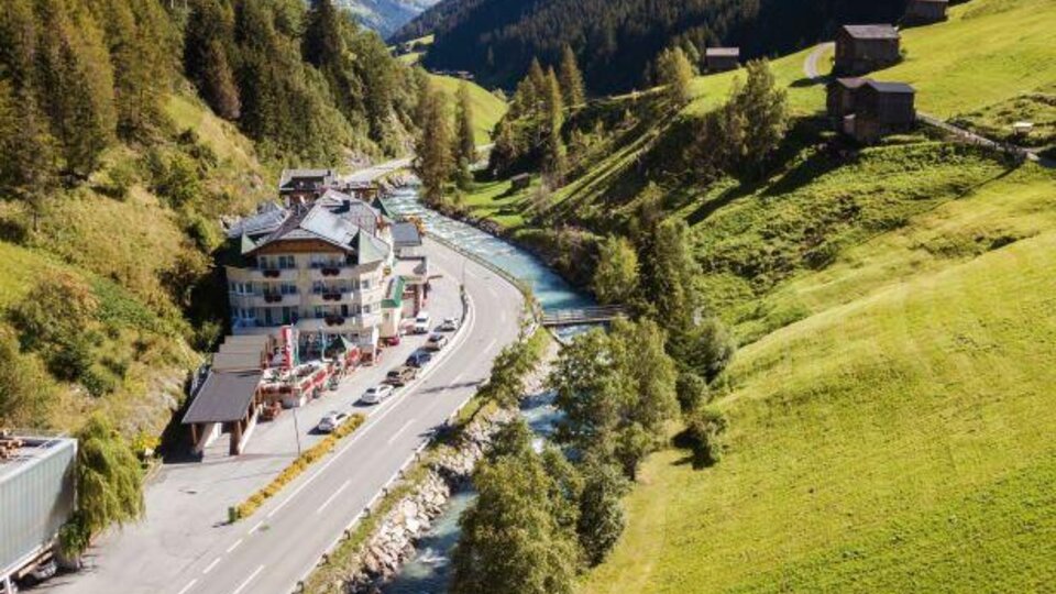 A picturesque landscape with a river flowing next to a road. In the background, green hills and some chalets can be seen.