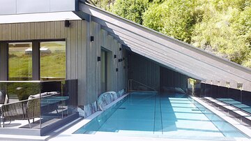 A modern house with a stylish pool and large windows. The covered terrace offers a view of the green surroundings.