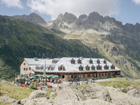 A cozy mountain cabin with outdoor seating and many guests. The surroundings are surrounded by impressive mountains and a clear blue sky.