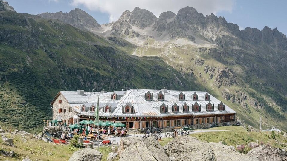 A cozy mountain cabin with outdoor seating and many guests. The surroundings are surrounded by impressive mountains and a clear blue sky.