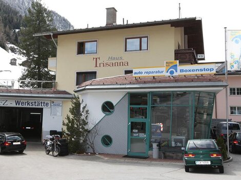Ein gemütliches Gebäude mit der Aufschrift "Haus Trisanna" und einer Werkstatt für Auto-Reparaturen. Im Vordergrund sind einige Autos und Fahrräder zu sehen.