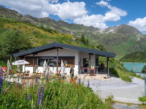 A modern building at the foot of the mountains, surrounded by green meadows and colorful flowers. In the background, majestic mountains and a clear blue sky can be seen.