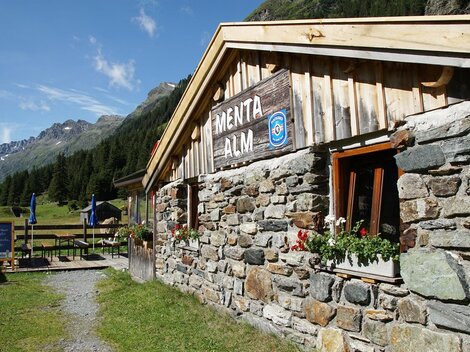 A cozy stone mountain hut in a picturesque mountain landscape. The surroundings are green, with mountains in the background and blue sky.