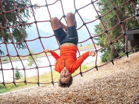 A boy is hanging upside down in a red climbing net and is happily looking at the camera. In the background, there are trees, a lake, and the blurred mountain landscape. The image captures a playful moment full of energy, courage, and lightness in the alpine outdoor experience.