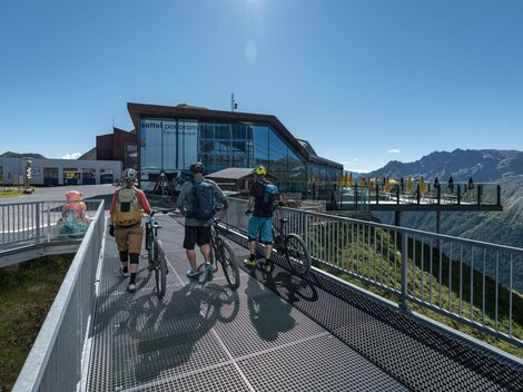 Three cyclists are standing on an observation platform overlooking the mountains. In the background, a modern building can be seen.