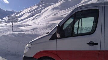 Ein Transporter steht vor einer verschneiten Landschaft. Im Hintergrund sind schneebedeckte Berge und ein Straßenschild sichtbar.