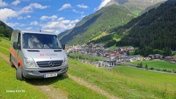 Ein Van steht auf einer Wiese mit Blick auf ein malerisches Dorf inmitten der Berge. Der Himmel ist blau mit einigen Wolken.