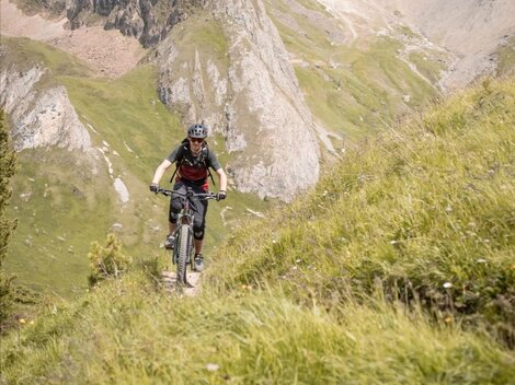 A cyclist rides on a narrow path through the green mountainous landscape. In the background, rocky mountains and a clear sky can be seen.